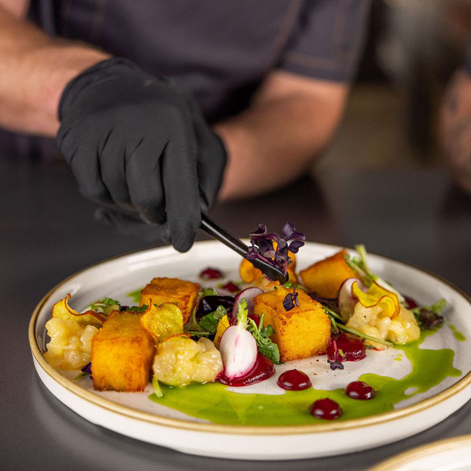 Koch mit Handschuhen richtet ein farbenfrohes Gericht auf weißem Teller an